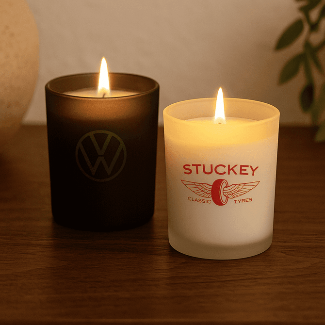 Two candles on a wooden surface: one black and one white, both with lit flames. The white candle has a logo.