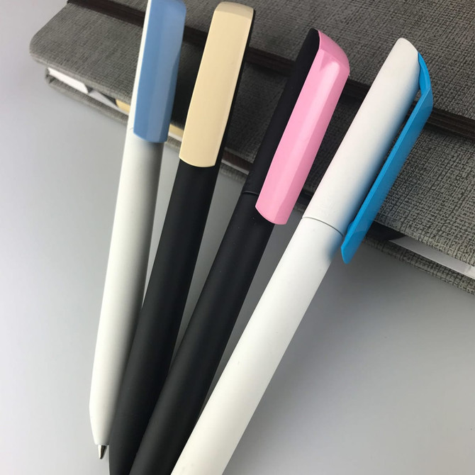 Four plastic pens in white, black, light blue, and pink, arranged on a grey notebook. The pens feature different cap designs and a logo.