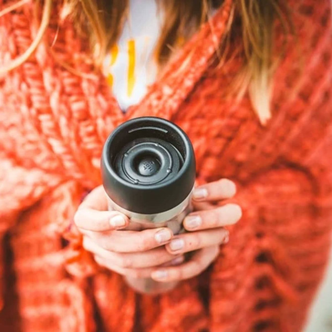 A travel mug with a stainless steel body and a black lid, held by a person wearing an orange knitted sweater.