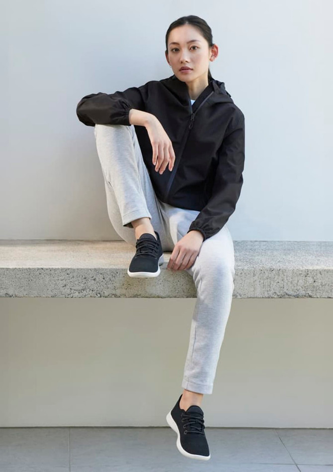 A woman sits casually wearing a black rain jacket, grey sweatpants, and black sneakers, against a light background.