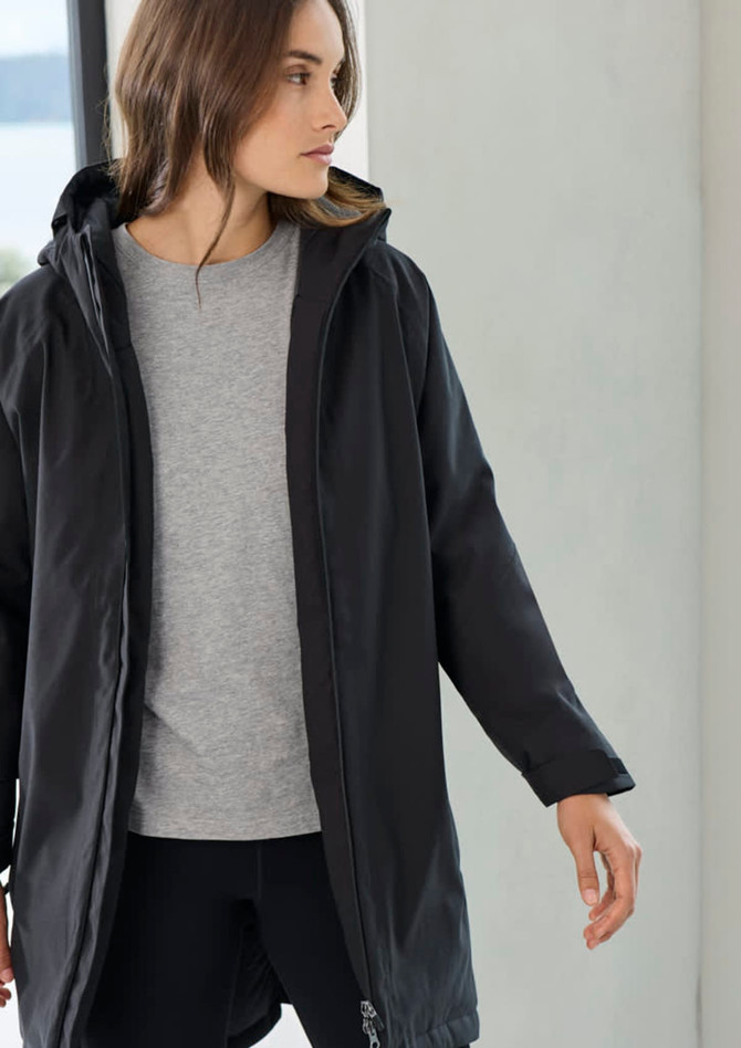 A unisex black rain jacket with a hood, worn over a grey top, showcased in a light-filled setting.