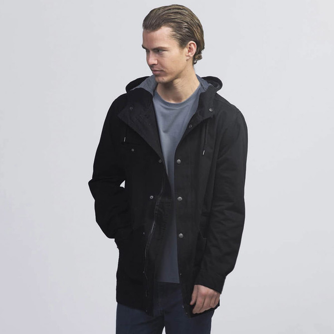 A black twill jacket featuring a hood and two front pockets, worn over a grey shirt by a model.