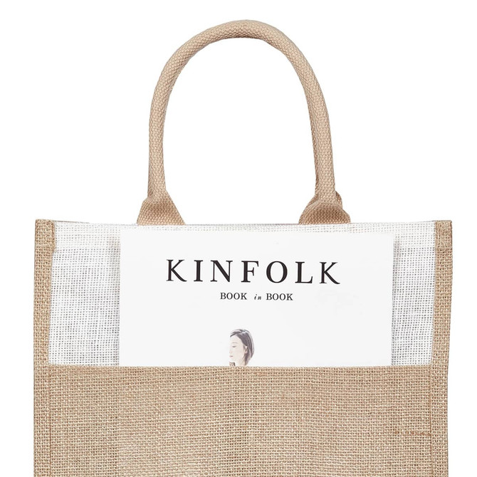 A jute bag in natural brown with handles, featuring a magazine partially visible in the front pocket and a logo.