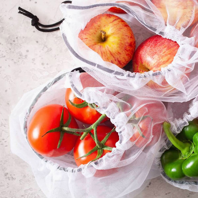 Nylon mesh produce bags containing red apples, ripe tomatoes, and green bell peppers on a textured surface.