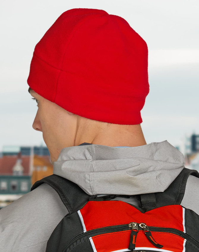 A red fleece beanie worn by a person with a grey hoodie and a black backpack. The background features a cityscape.
