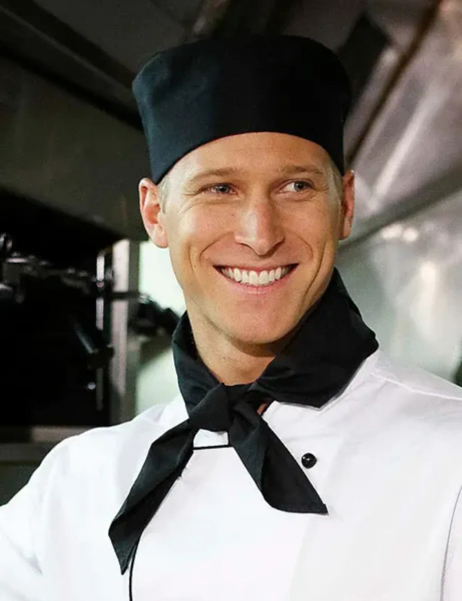A chef wearing a black scarf and black cap, smiling in a kitchen setting. The scarf complements his white chef's uniform.