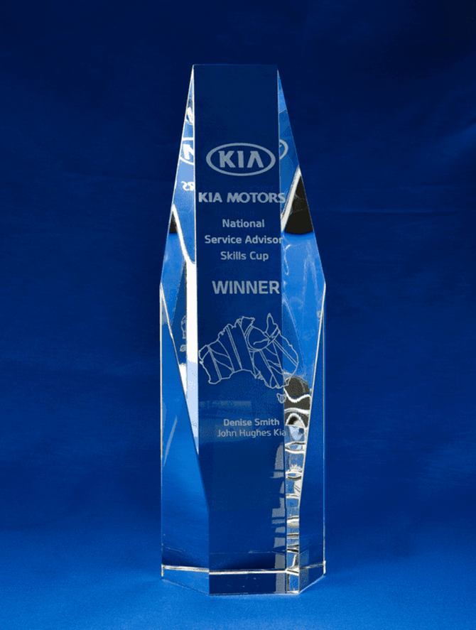 A large, clear crystal trophy in a hexagonal shape, featuring engraving and a logo. The background is blue.