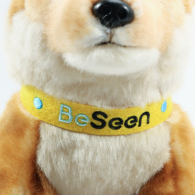 A plush dingo toy in brown with a yellow collar featuring "Be Seen" in black and blue embroidery.