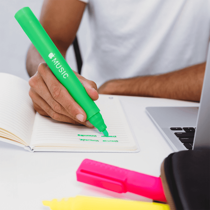A hand holding a bright green jumbo highlighter above a notebook, with pink and yellow highlighters visible nearby.