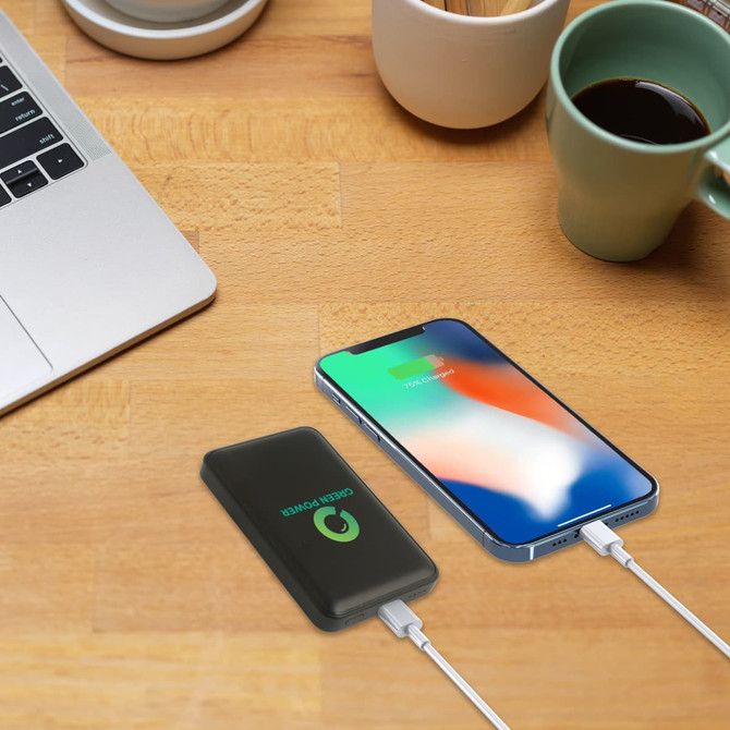 A black 10000mAh power bank with a logo beside a smartphone, on a wooden desk with a coffee cup.