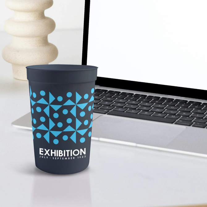 A plastic stadium cup in black featuring blue geometric designs, placed next to a laptop on a white desk.