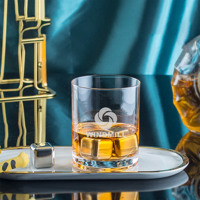 A whiskey glass with a logo, containing amber liquid and ice cubes, placed on a sleek tray near golden decor.