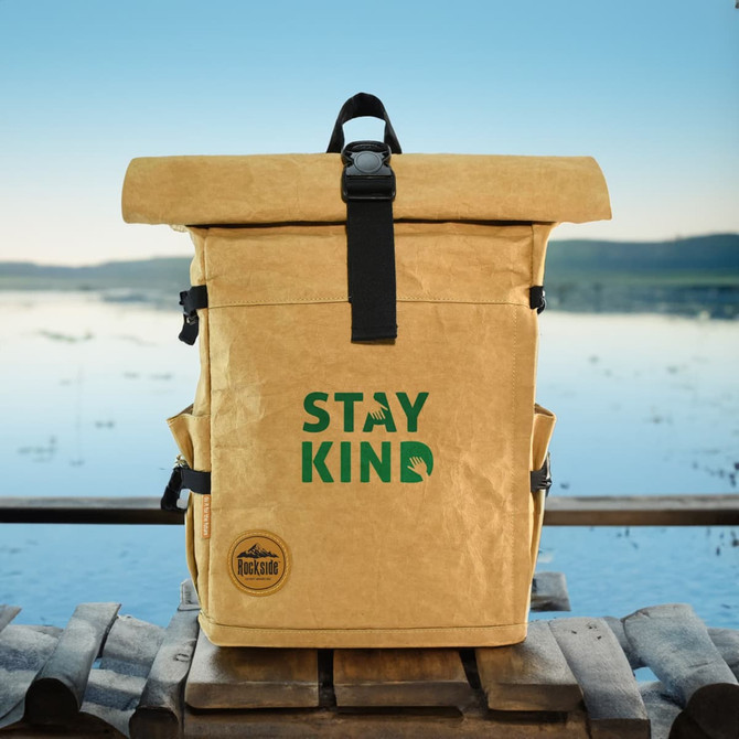 A yellow kraft paper laptop backpack with a roll-top design and a logo on the front, set against a scenic lake backdrop.