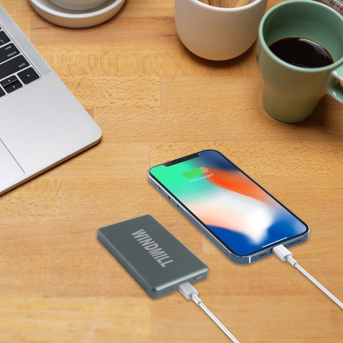 A grey aluminium power bank sits on a wooden table next to a smartphone, connected by a charging cable.