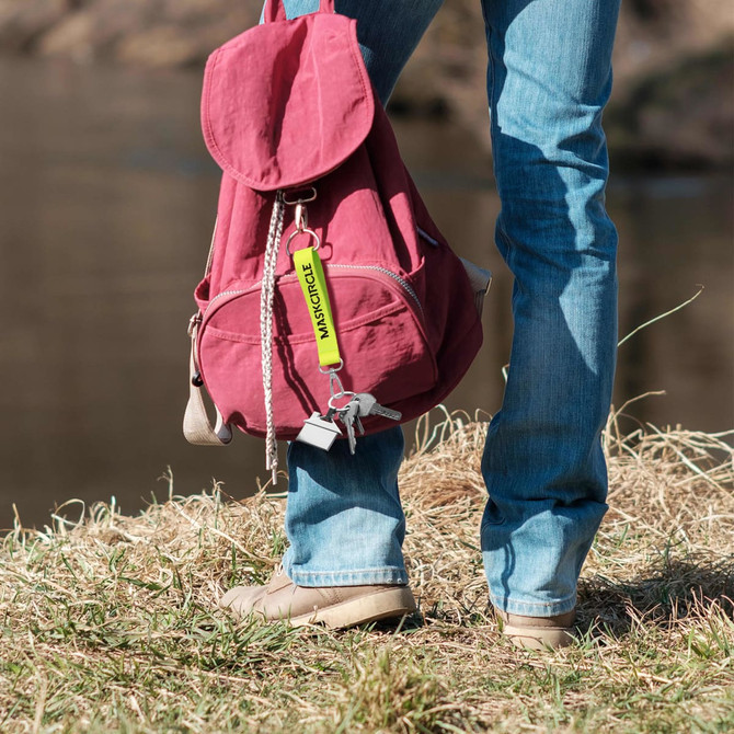 A keyring in yellow attached to a maroon backpack, held by a person standing on grass near a riverbank.