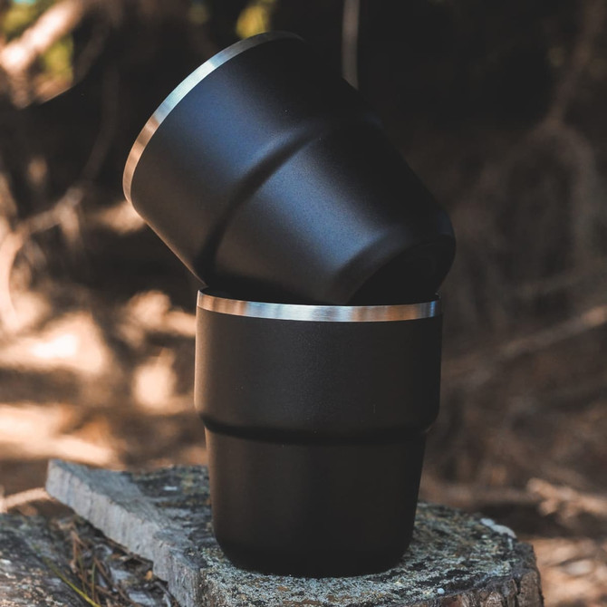 A set of two stackable cups in black with a silver rim, placed on a rustic wooden surface.