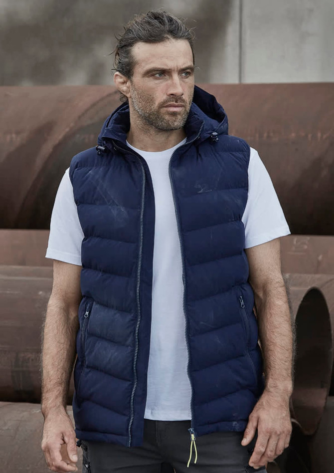A man wearing a navy blue unisex hooded puffer vest over a white t-shirt, standing in an industrial setting.