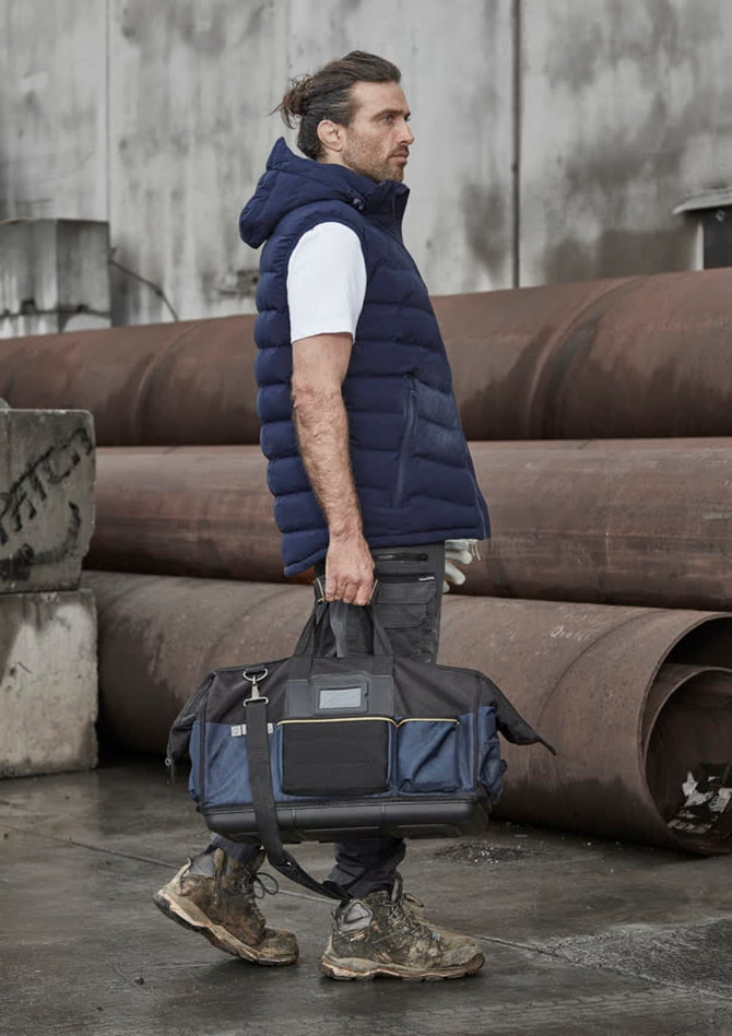 A man wearing a navy hooded puffer vest holds a black and navy duffle bag, standing against industrial backdrops.