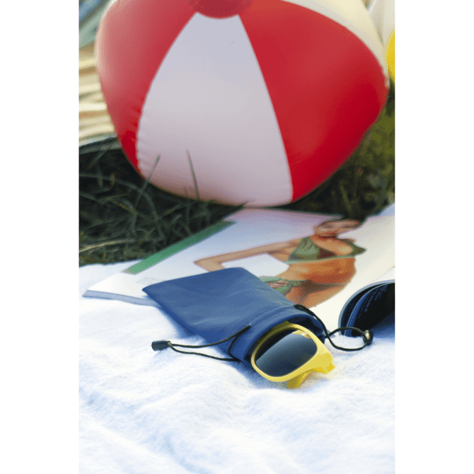 A navy pouch with sunglasses, placed on a white towel beside a beach ball and a magazine. The pouch has a drawstring closure.