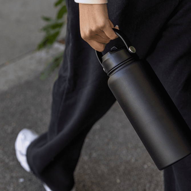 A drink bottle in matte black, held by a person, featuring a handle and a lid, with a logo on the side.