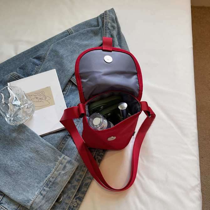 A red canvas shoulder bag with a grey interior, containing a drink bottle and other items, sits on a bed.