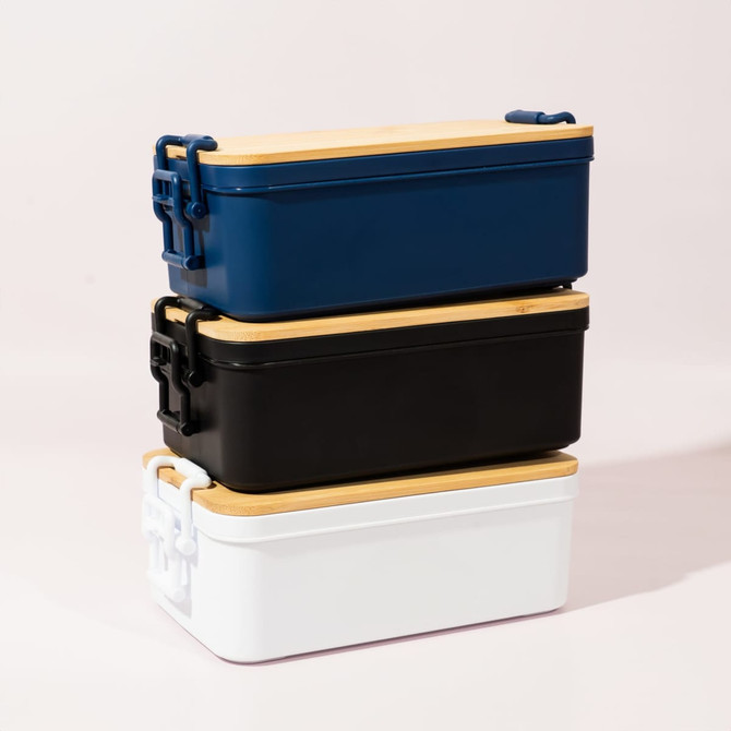 Three stacked lunch boxes in navy blue, black, and white, each with a wooden lid and a branded logo.