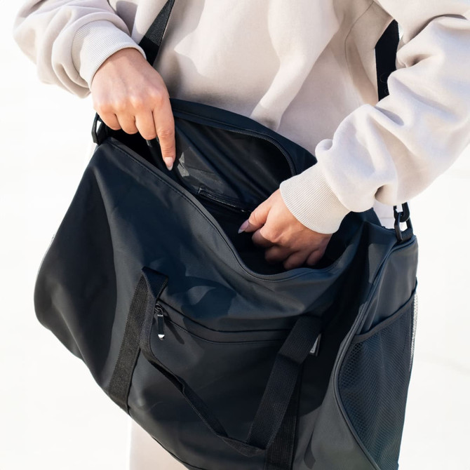 A black RPET duffle bag with multiple compartments, being held by a person. The bag has a shoulder strap and a logo.