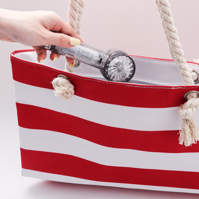A hand holds a transparent cooling fan as it is placed inside a red and white striped tote bag.