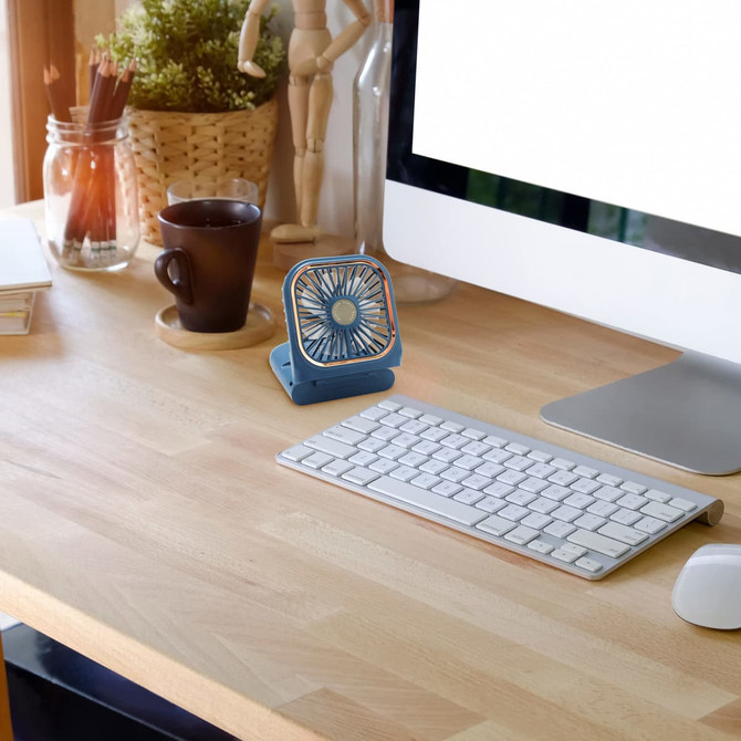 A blue folding neck fan with a phone stand rests on a wooden desk beside a cup and a computer keyboard.
