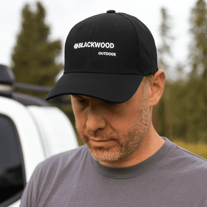 A black strapback baseball cap with a logo, worn by a person outdoors near a vehicle.