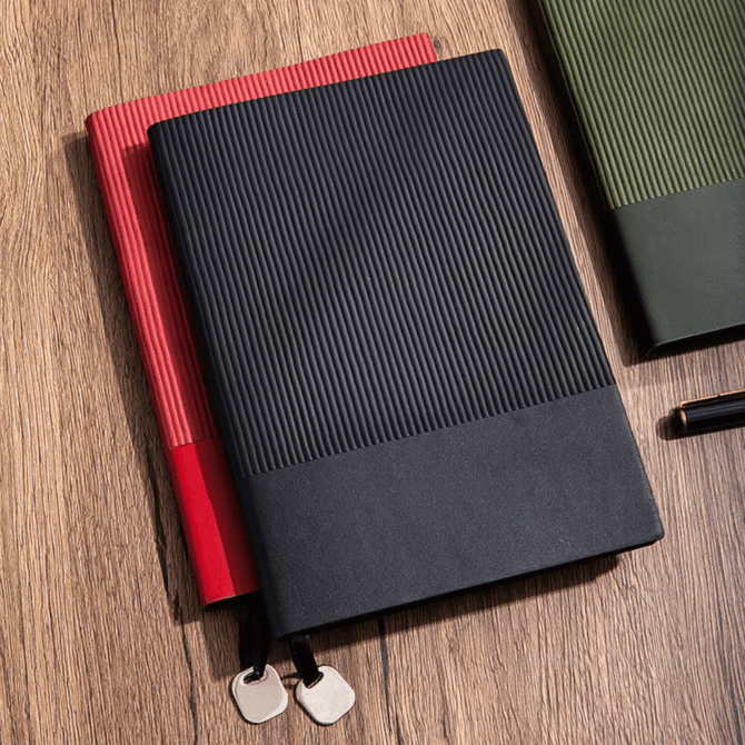 A black ribbed leather notebook with a red notebook beside it on a wooden surface. The notebook has branding and two silver tags.