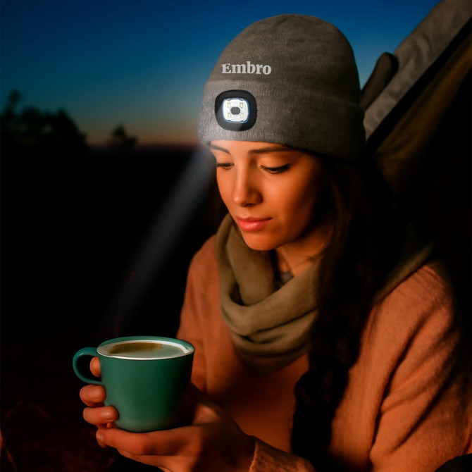 A grey beanie with a light, worn by a woman holding a green cup, against a twilight background.