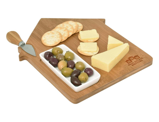 A wooden cheese board shaped like a house, featuring cheese slices, crackers, olives, and a cheese knife.