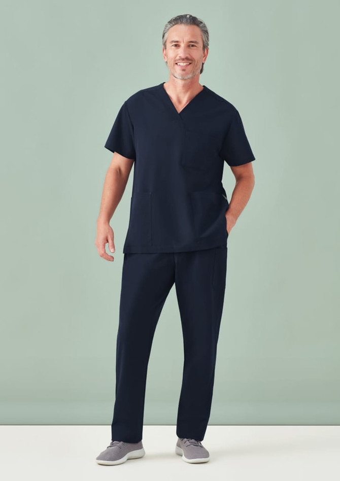A man wearing a unisex navy V-neck scrub top and matching pants, standing against a light green background.