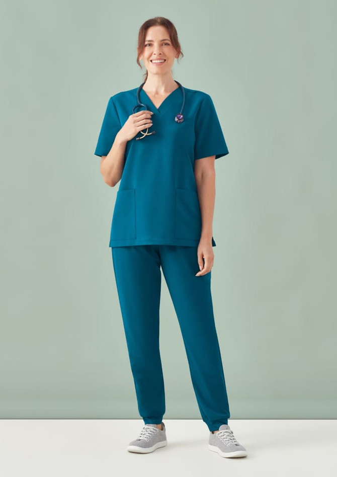 A woman models a teal women's scrub top with a V-neck and matching scrub pants, featuring a logo.