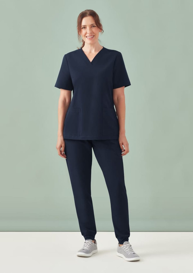 A woman wearing navy women's jogger scrub pants and a matching scrub top, standing against a muted background.