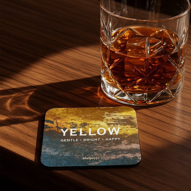 A square frosted metal coaster in yellow and blue, placed beside a glass of whiskey on a wooden surface.