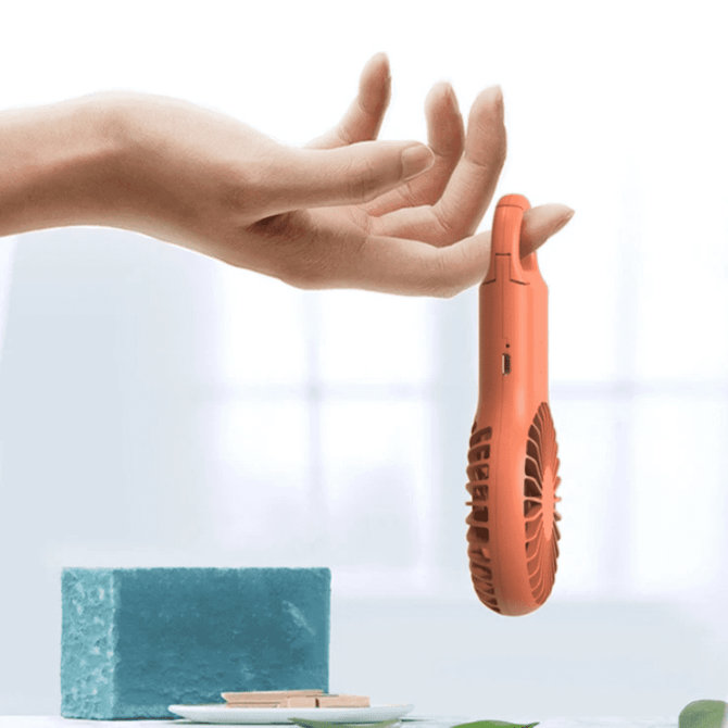 A handheld, coral pocket fan is suspended above a blue sponge and a dish, with a soft background.