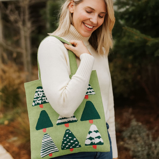 A green tote bag featuring a pattern of Christmas trees, held by a smiling woman in a white sweater.