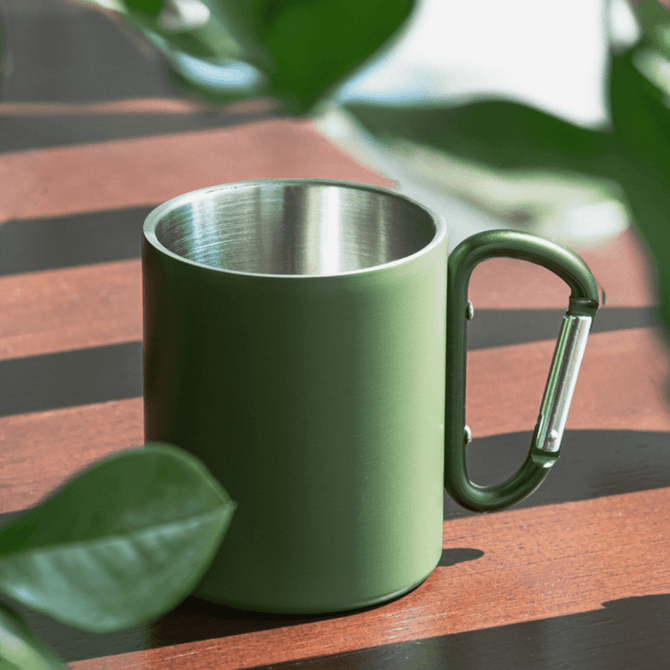 A green thermo mug with a stainless steel interior and a carabiner-style handle, resting on a wooden surface.