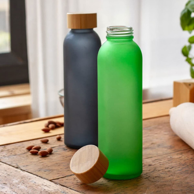 A drink bottle in frosted green glass with a wooden lid, next to a frosted grey bottle, on a wooden surface.