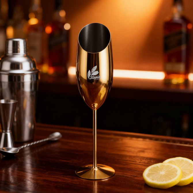 A stainless steel slanted champagne flute in gold colour, featuring a logo, on a wooden bar with bar tools and lemons.