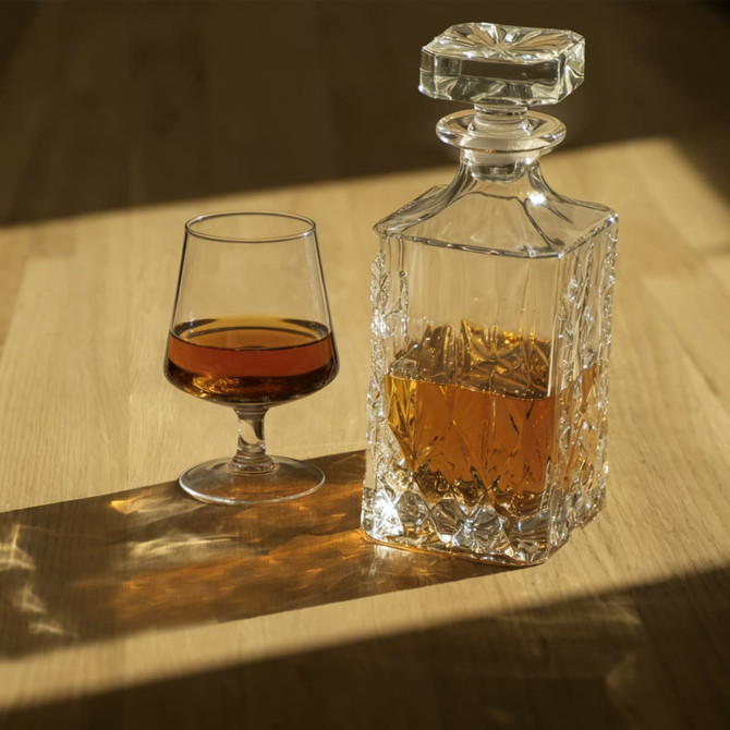 A crystal decanter and a snifter glass filled with amber liquid sit on a wooden surface, casting soft shadows.