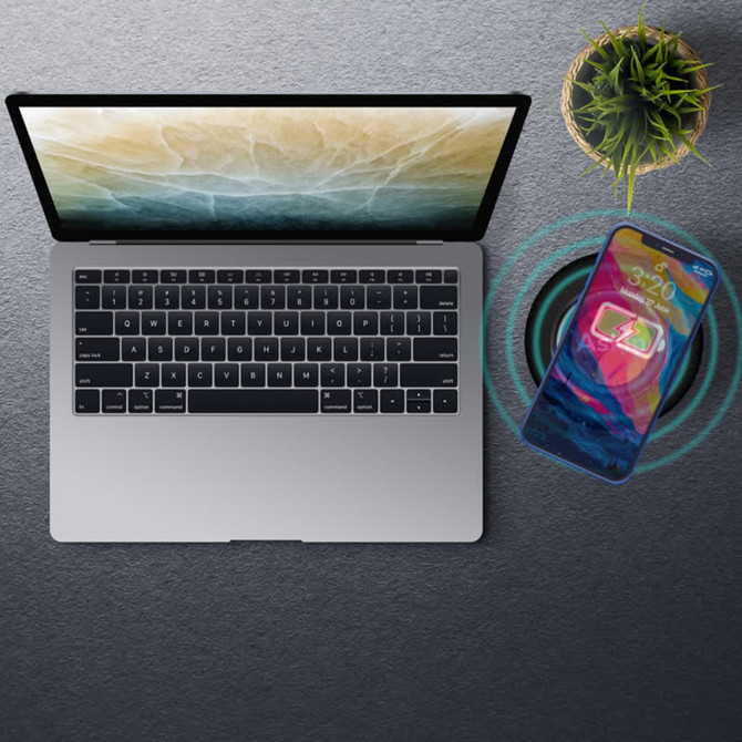 A silver laptop is placed on a textured surface next to a phone wirelessly charging. A small plant is also visible.