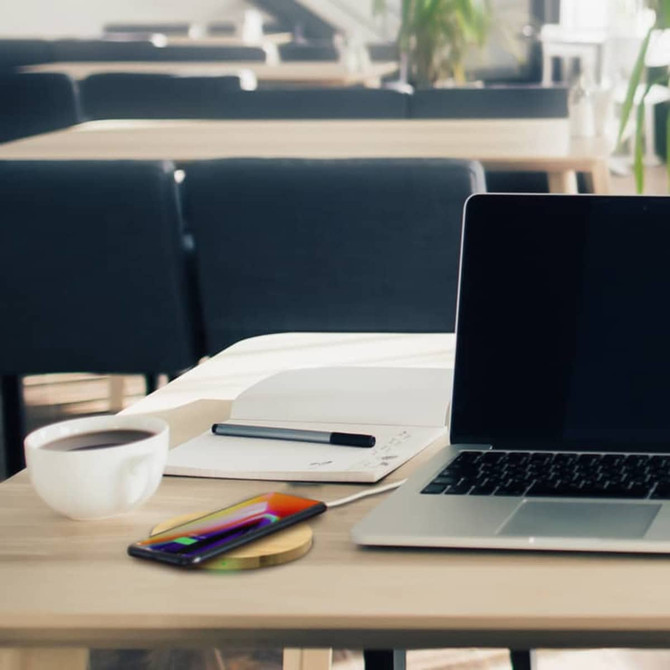 A wireless bamboo fast charger in a natural wood tone on a table near a laptop, notebook, pen, and coffee cup.