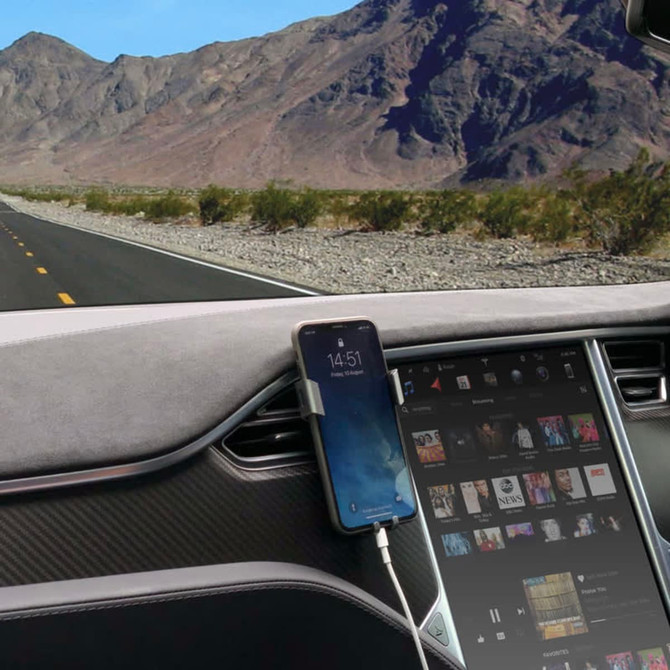 A car phone holder in silver, securely holding a smartphone on a dashboard, with a scenic view of a highway and mountains.