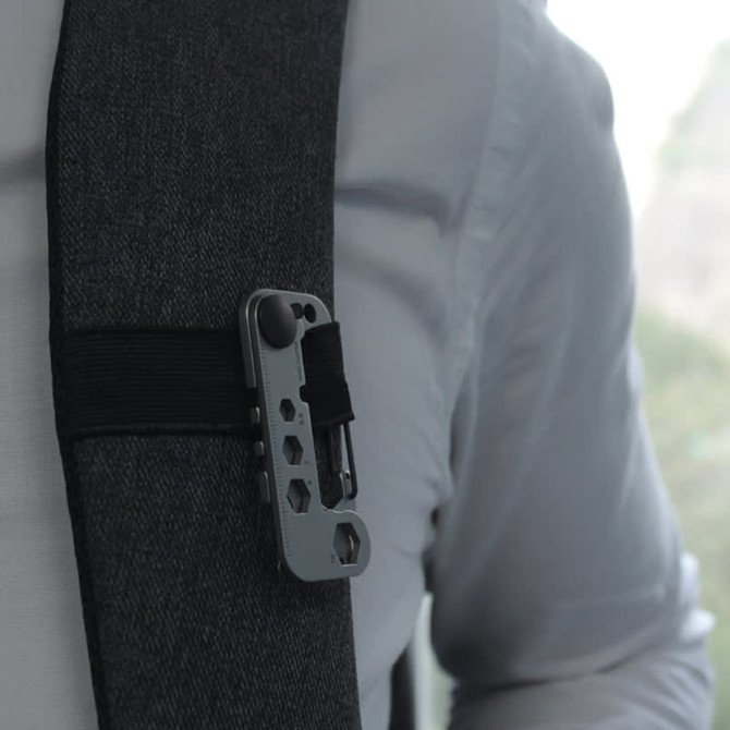 A grey multi-tool clipped onto a strap, featuring various tool functions and a branding logo.