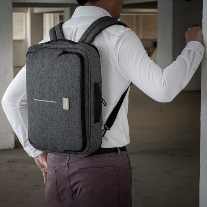 A hybrid laptop bag in grey, worn on the back of a person, featuring a minimalist design and a logo.