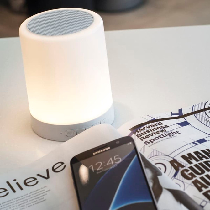 A white touch lamp speaker with a soft glow sits on a table next to a smartphone and a magazine.