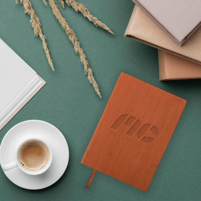 A5 PU leather notebook in brown with a logo, surrounded by stack of notebooks and a coffee cup on green background.
